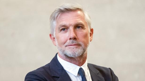 Ein Mann mit grauem Haar und Bart trägt einen dunklen Anzug.