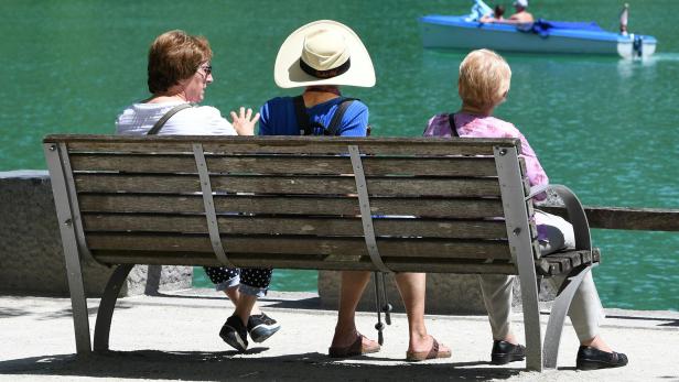 Drei Personen sitzen auf einer Bank mit Blick auf einen See, auf dem ein Boot fährt.