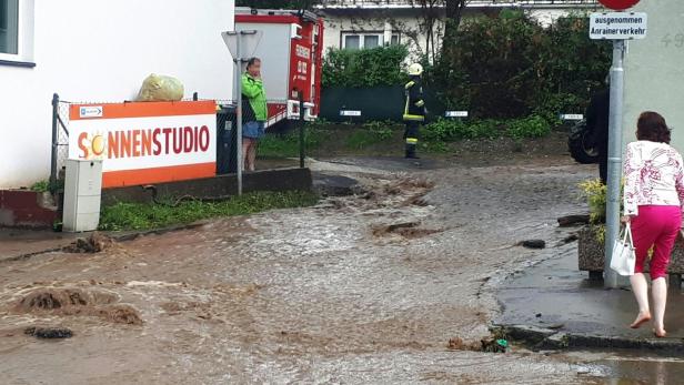 Überschwemmte Straße mit Feuerwehrleuten und einer Frau, die barfuß durch das Wasser geht.