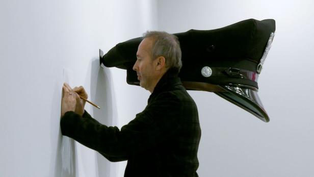 Ein Mann zeichnet an einer Wand neben einer übergroßen Polizeiuniformmütze.