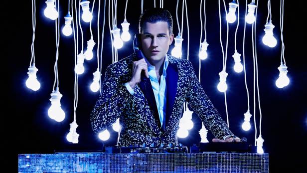 Ein DJ mit gemustertem Sakko steht vor einem Mischpult, umgeben von Glühbirnen.