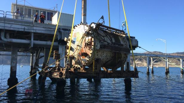 Ein großes, rostiges Objekt wird mit einem Kran vom Wasser gehoben.