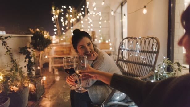 Eine Frau prostet mit einem Glas Rotwein auf einer mit Lichterketten dekorierten Terrasse zu.