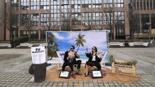 Vor dem EU-Parlament sitzen zwei Personen mit Geld auf Liegestühlen vor einer Strandkulisse.