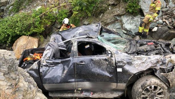 Ein schwer beschädigter schwarzer Pick-up liegt zwischen Felsen, während Rettungskräfte vor Ort sind.
