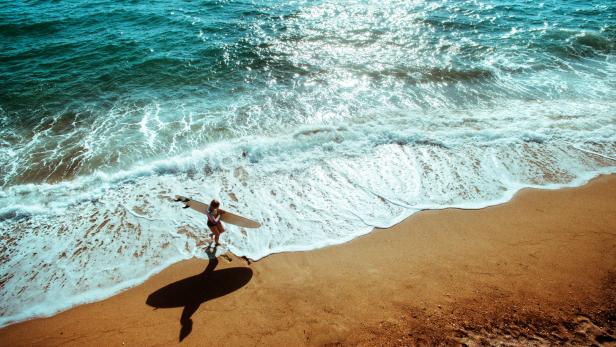 Eine Frau mit Surfbrett steht am Strand im türkisfarbenen Wasser.