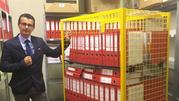 Ein Mann in einem Anzug steht neben einem gelben Rollwagen voller roter Aktenordner.