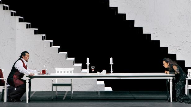 Eine Szene aus einer Opernaufführung mit zwei Sängern an einem langen Tisch vor einer Treppe.