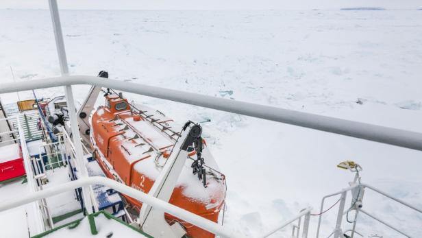 Ein orangefarbenes Rettungsboot auf einem Schiff, bedeckt mit Schnee, vor einer eisigen Landschaft.