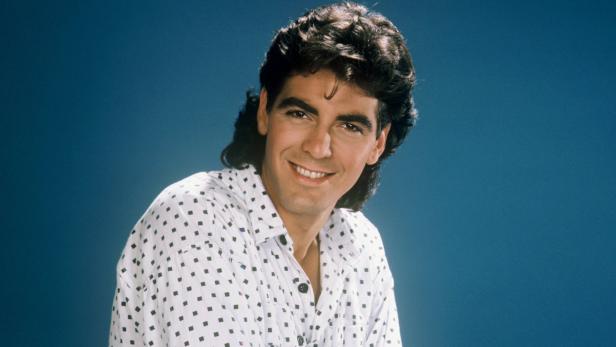 Ein junger George Clooney lächelt in einem weiß gemusterten Hemd vor blauem Hintergrund.