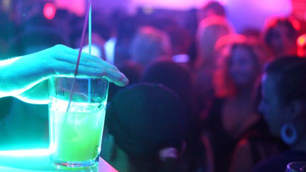 Ein Trinkglas mit einer Hand darauf, im Hintergrund eine Szene aus einem Tanzklub