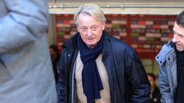 Ein älterer Mann mit Schal und Jacke steht im Vordergrund.