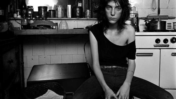 Schwarzweißporträt von Patti Smith, die in einer Küche sitzt und ein Glas hält.