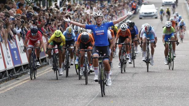 Eine Radrennfahrerin überquert mit erhobenen Armen die Ziellinie, gefolgt von anderen Fahrerinnen.