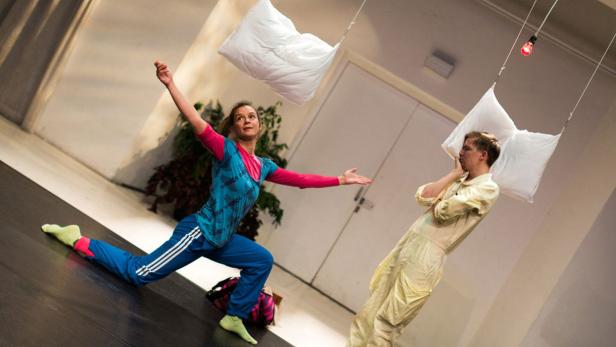 Eine Frau in Sportkleidung und ein Mann in einem hellen Overall interagieren auf einer Bühne mit hängenden Kissen.