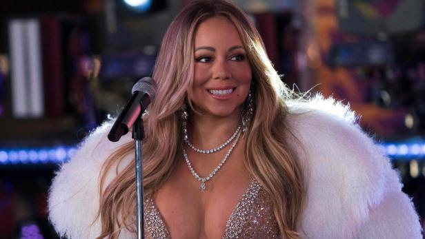 Mariah Carey steht lächelnd mit Glitzerkleid und weißem Fellmantel vor einem Mikrofonständer