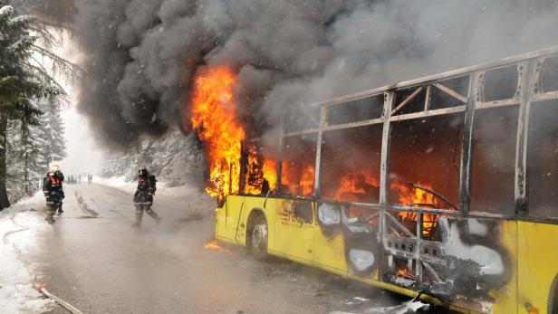 Ein gelber Bus brennt auf einer schneebedeckten Straße, während Feuerwehrleute im Hintergrund stehen.