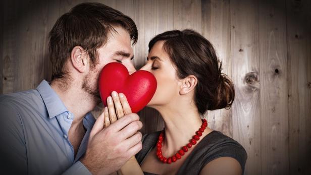 Ein Paar hält ein rotes Herz vor ihre Gesichter, als ob sie sich küssen würden.
