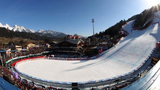 Panoramaaufnahme einer Skipiste mit Zuschauern und schneebedeckten Bergen im Hintergrund.