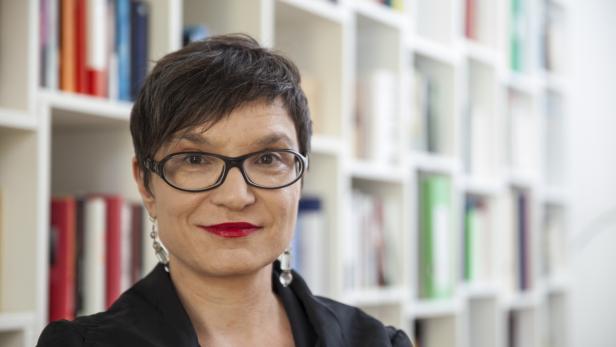 Eine Frau mit Brille und rotem Lippenstift vor einem Bücherregal.