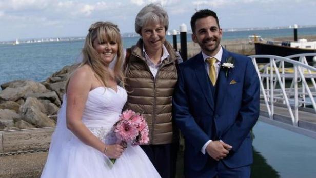 Eine Braut, ein Bräutigam und Theresa May posieren für ein Foto am Meer.