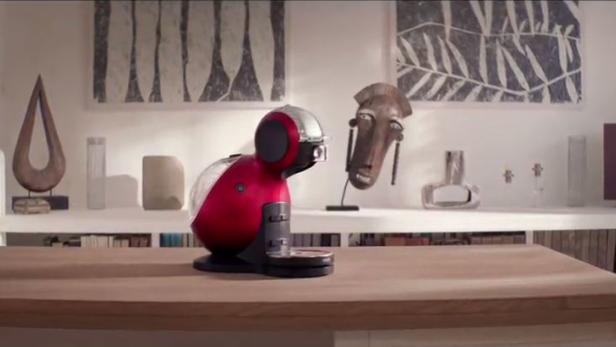 Eine rote Kaffeemaschine steht auf einem Holztisch vor einem Regal mit Kunstgegenständen.