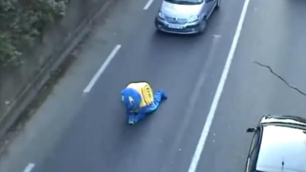 Eine Person im Schneckenkostüm kriecht auf einer Straße mit Autos.