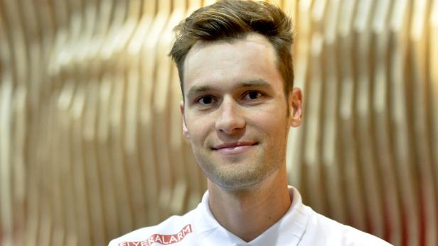 Ein lächelnder Mann mit braunen Haaren und einem weißen Poloshirt vor einem gemusterten Hintergrund.