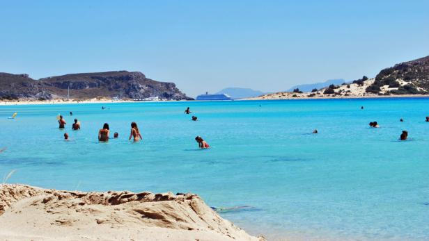 Menschen schwimmen im türkisfarbenen Wasser an einem sonnigen Strand.