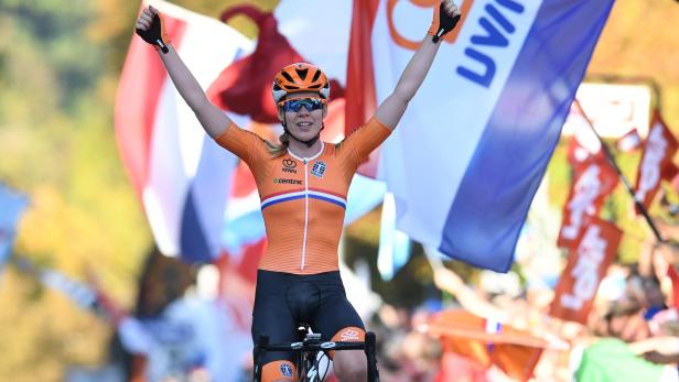 Eine Radrennfahrerin jubelt mit erhobenen Armen vor niederländischen Flaggen.