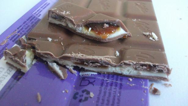Eine teilweise aufgebrochene Schokoladentafel mit Karamellfüllung liegt auf einer lila Verpackung.
