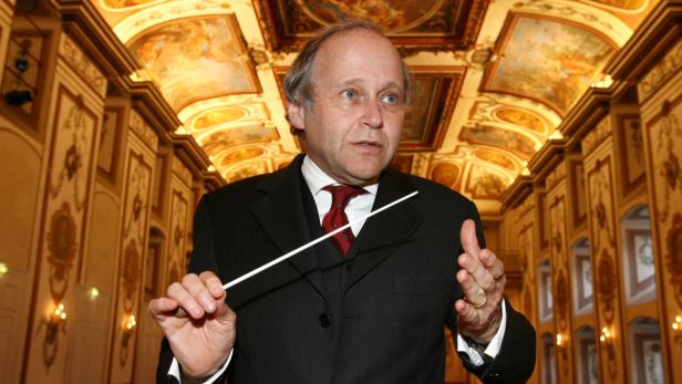 Ein Dirigent mit Taktstock vor einem reich verzierten Saal.