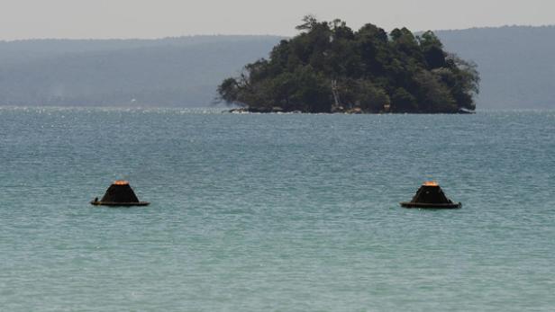 Zwei brennende Flöße treiben auf dem Wasser vor einer bewaldeten Insel.