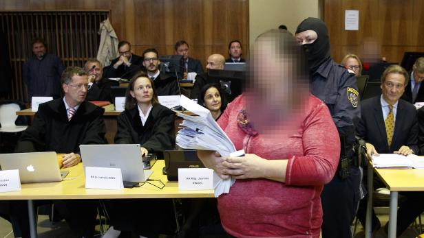 Eine verschleierte Frau mit einem Stapel Papier wird von einem Justizbeamten in einem Gerichtssaal bewacht.