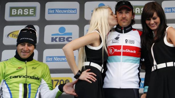 Ein Radrennfahrer wird auf dem Podium von zwei Frauen flankiert und von einer geküsst.