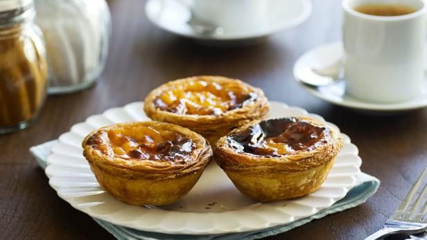 Drei Pastéis de Nata auf einem Teller mit Tassen im Hintergrund.