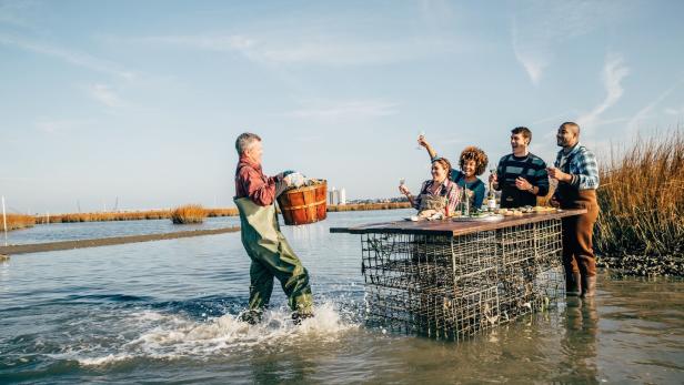 Ein Mann watet mit einem Eimer voller Austern zu einer Gruppe, die an einem Tisch im Wasser steht und Wein trinkt.