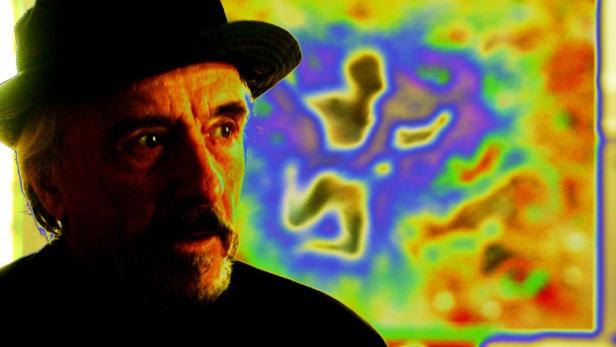 Ein Mann mit Hut blickt vor einem farbenfrohen, abstrakten Gemälde nach rechts.