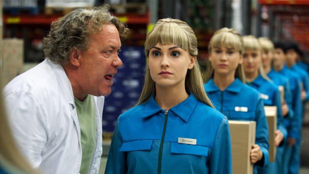 Ein Mann in einem Laborkittel spricht eine junge Frau namens Hermione an, die mit anderen Frauen in blauen Overalls in einer Reihe steht.