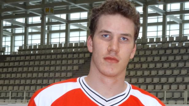 Ein junger Mann mit blauen Augen und einem orangefarbenen Trikot steht in einer Sporthalle.