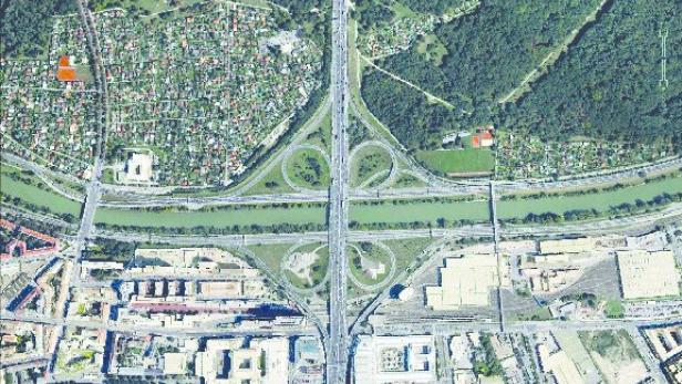 Eine Luftaufnahme einer Autobahnkreuzung über einem Fluss, umgeben von Stadt, Wald und Kleingartensiedlungen.