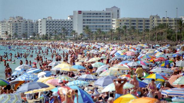 Ein überfüllter Strand mit vielen Menschen und bunten Sonnenschirmen vor großen Hotels.