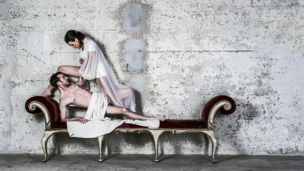 Ein Mann liegt auf einer roten Chaiselongue, während eine Frau in einem weißen Kleid über ihm steht.
