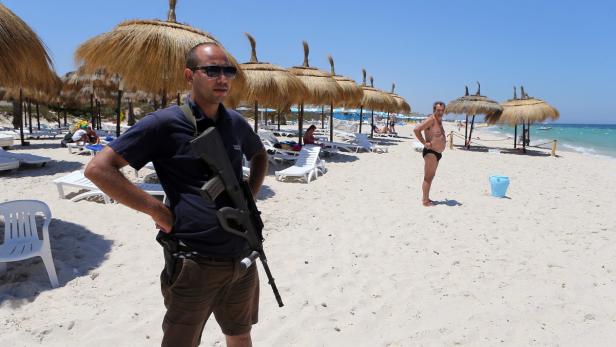 Ein bewaffneter Mann steht an einem Strand mit Sonnenschirmen und Badegästen.