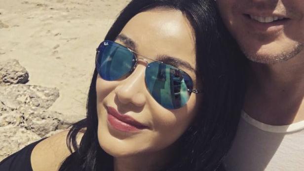 Eine Frau mit Sonnenbrille und ein Mann posieren am Strand für ein Selfie.