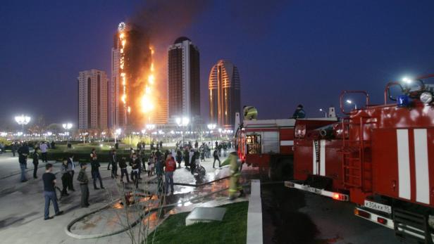 Ein Hochhaus steht in Flammen, während Feuerwehrleute und eine Menschenmenge zusehen.