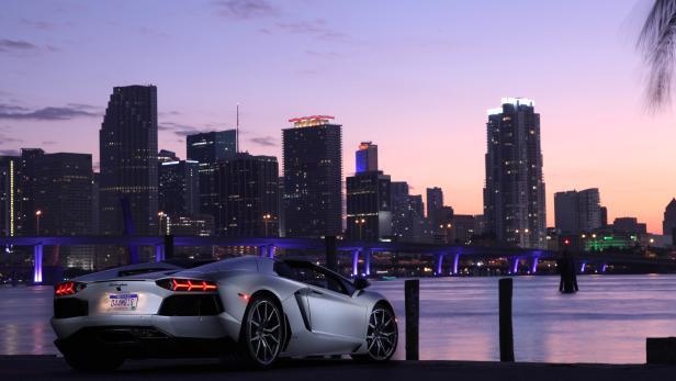 Ein silberner Lamborghini vor der Skyline von Miami bei Sonnenuntergang.
