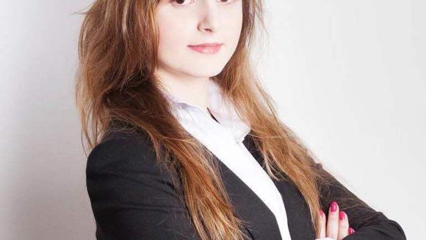 Eine junge Frau mit braunem Haar und Blazer steht mit verschränkten Armen da.