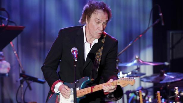 Ein Mann in einem schwarzen Anzug spielt auf einer Bühne Gitarre.