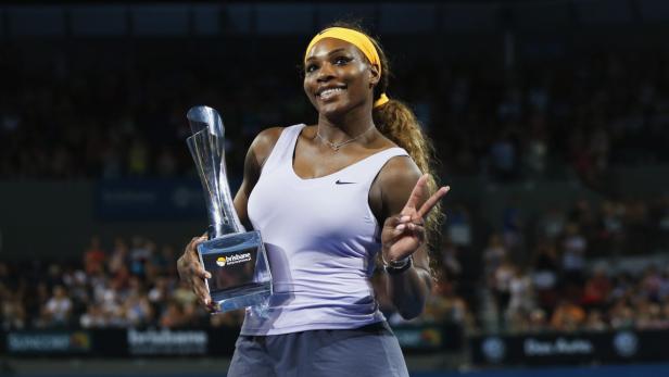 Serena Williams hält eine Trophäe in der Hand und zeigt ein Victory-Zeichen.